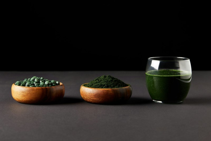 glass of spirulina smoothie, bowls with spirulina powder and spirulina pills on black background